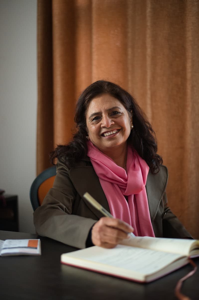 Notre responsable pays pour le Népal, Nirmala Sharma. Elle est assise à son bureau et écrit quelque chose dans un carnet. Elle a de longs cheveux noirs et porte une écharpe rose et un blazer gris-brun.