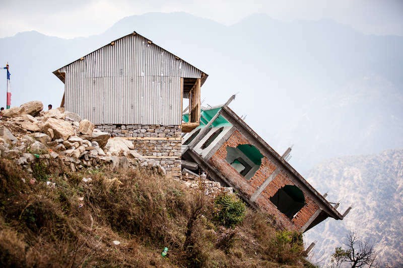 The picture shows a house that has slid down parts of a slope as a result of the earthquakes and is now standing at an angle on the slope. The walls are broken, the window panes outside.