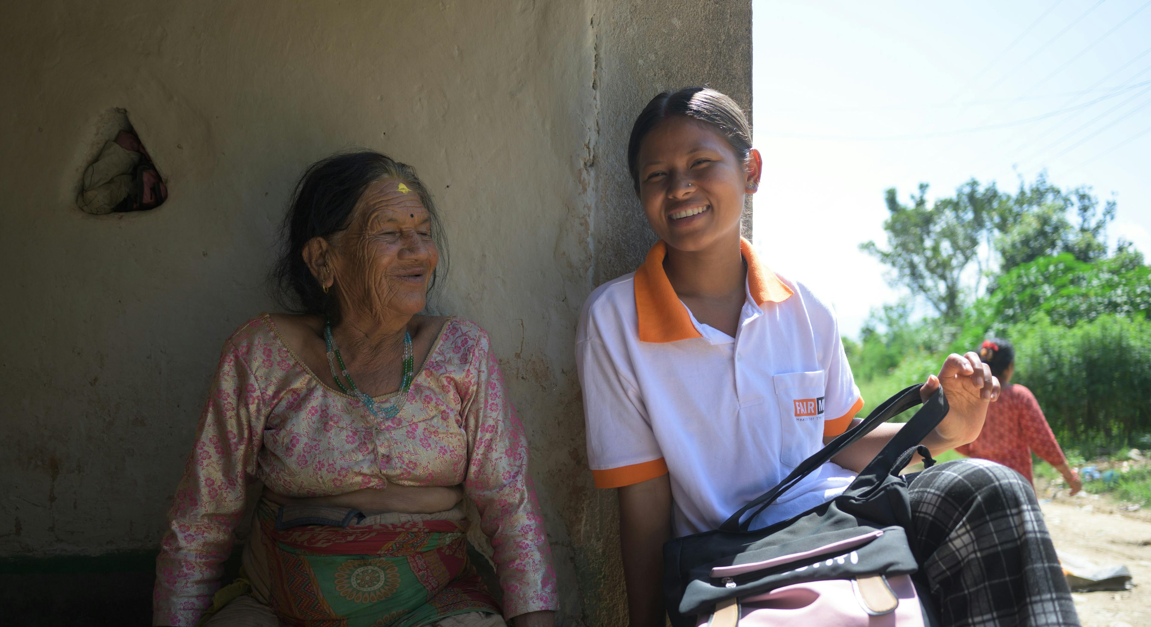 Sujita est assise par terre avec une femme âgée devant une maison et discute avec elle.