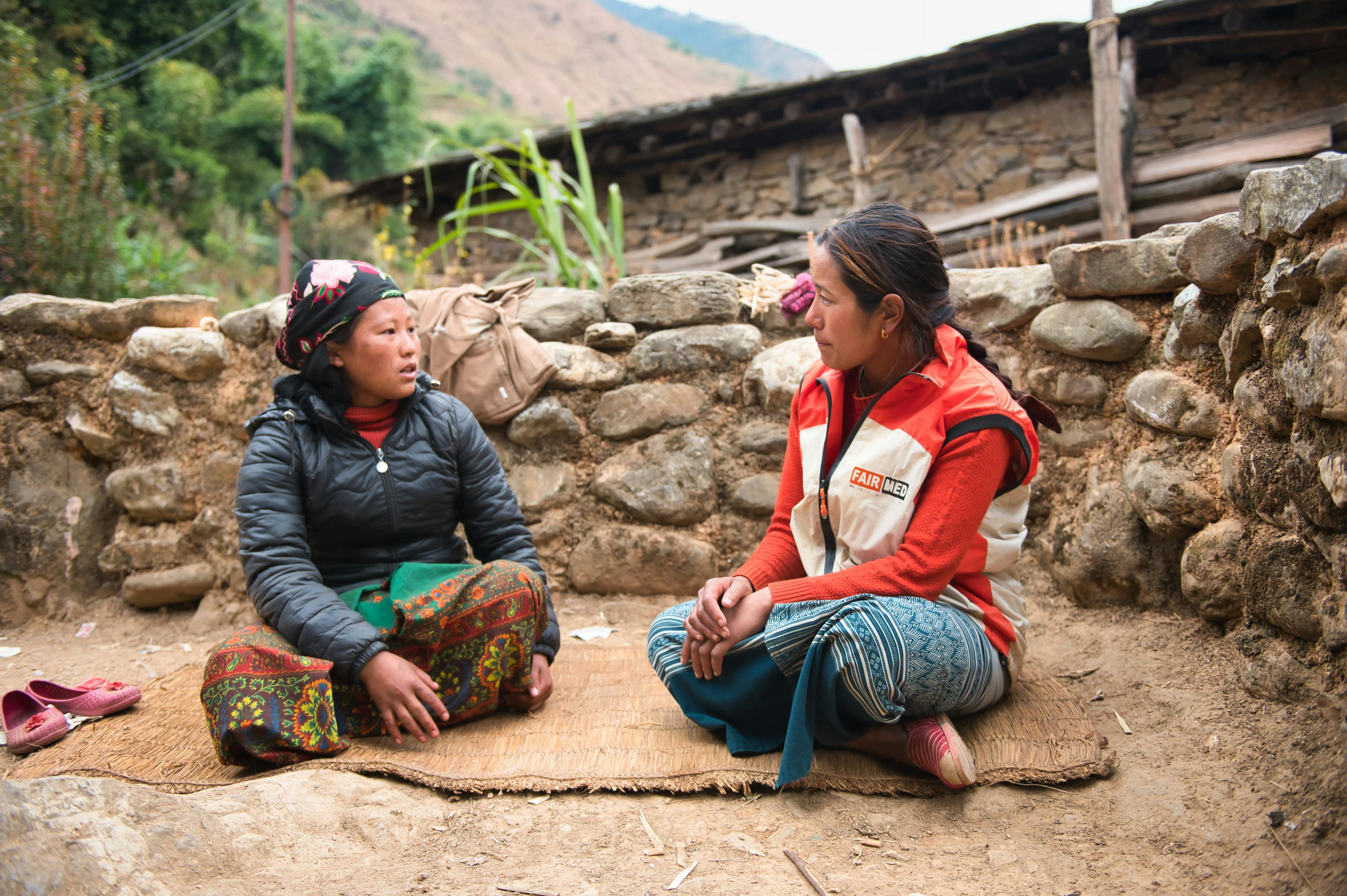 Gyan, jeune collaboratrice de FAIRMED, est assise par terre avec une jeune mère et lui donne des conseils sur sa santé et celle de son enfant. Elles sont assises sur une natte, avec en arrière-plan un mur de pierre et des éléments d'une hutte rudimentaire. La scène se déroule dans un village isolé de Baglung, au Népal.