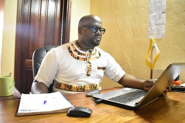 Notre responsable pays en République centrafricaine Jacques Christian Minyem. Il porte une chemise blanche avec des motifs traditionnels et des lunettes noires. Il est chauve et porte une barbe. Il est assis devant son ordinateur, à son bureau.