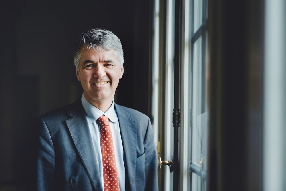 Alec von Graffenried steht vor einem schwarzen Hindergrund. Er trägt ein blaues Hemd, einen dunkelblauen Blazer und eine rote Krawatte mit weissen Punkten.