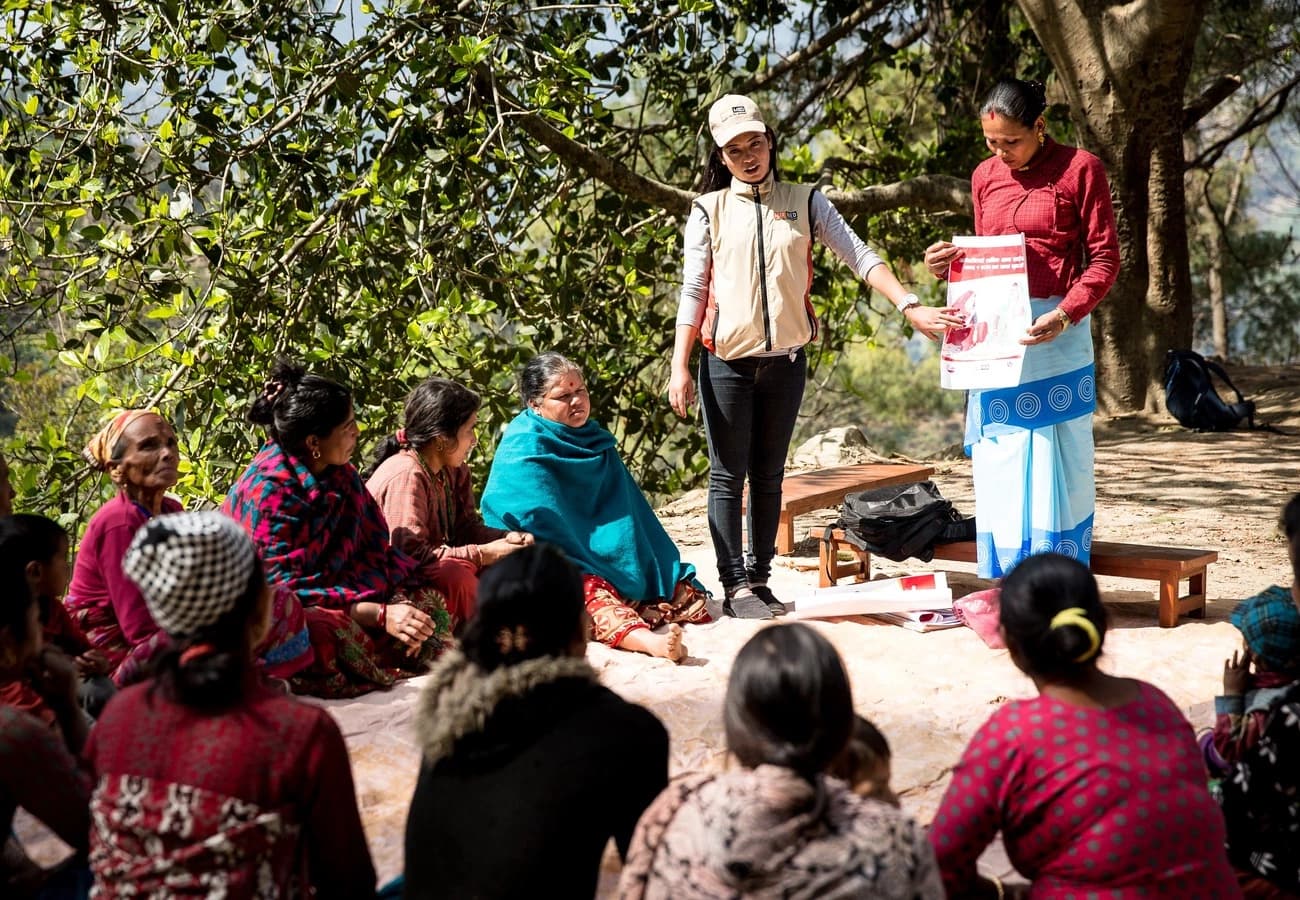 Unsere Mitarbeiterin Nobina leitet eine Müttergruppe. Sie steht und zeigt auf eine Informationsbroschüre, die eine der Mütter in der Hand hält, und erklärt den sitzenden Frauen etwas. Es sind rund zehn Frauen, die im Freien am Boden sitzen.