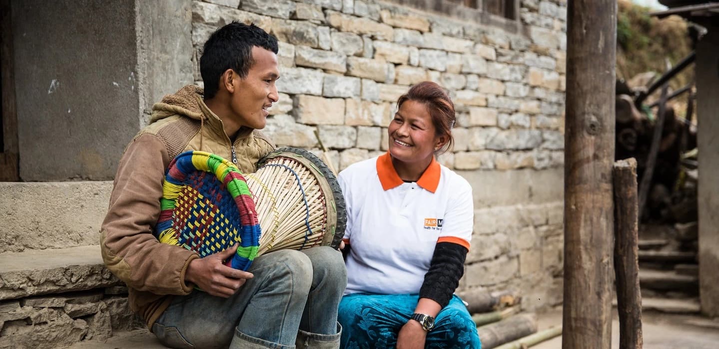 A FAIRMED employee in a white polo shirt with FAIRMED logo is sitting with a blind young man in front of the entrance of a rudimentary hut. She is advising him on health issues.