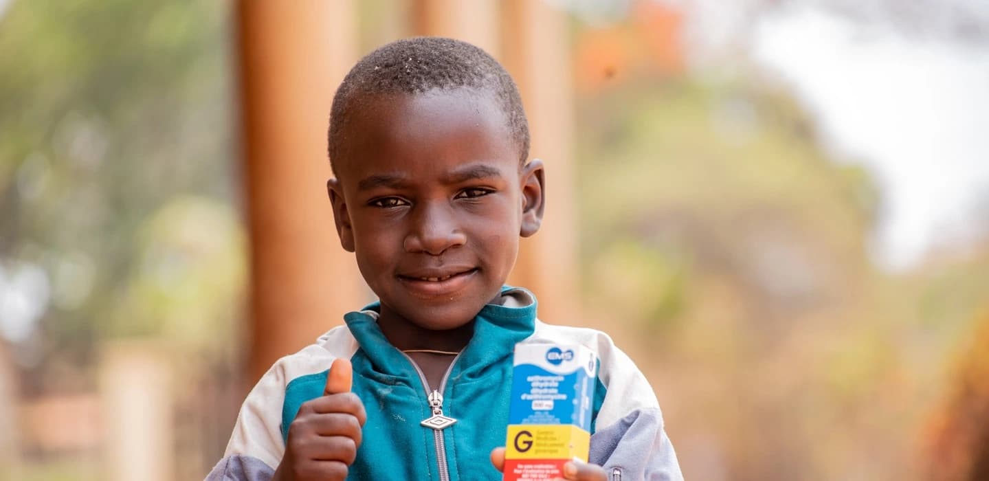 A young boy in Cameroon looks into the camera with his thumb outstretched and antibiotics in his hand. He received this from FAIRMED employees.