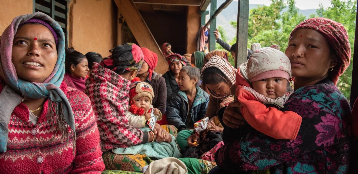 Auf dem Bild sind Mütter und ihre Kinder in Nepal zu sehen. Sie sitzen eng beieinander unter einem Dach. Sie sind winterlich und bunt gekleidet.