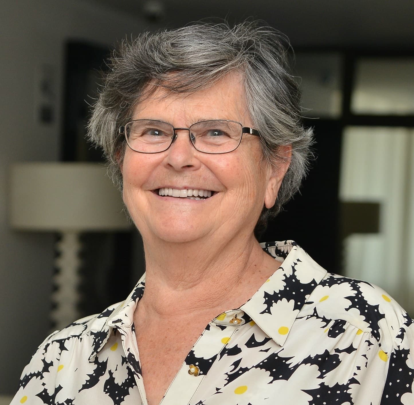 Ruth Dreifuss steht vor einem neutralen Hintergrund. Sie trägt eine silberfarbene Brille und eine schwarz-weisse Bluse.