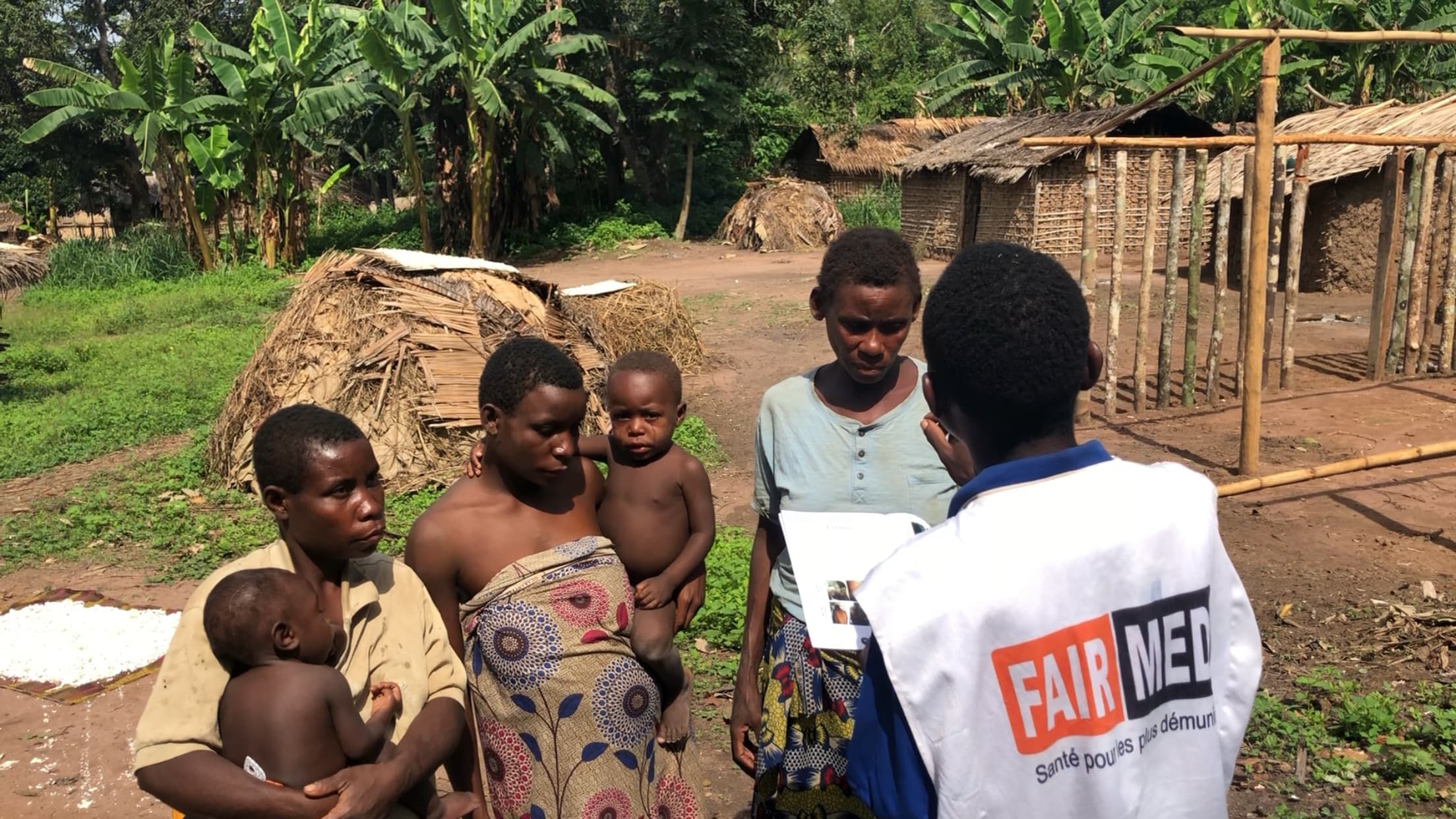 Un collaborateur de FAIRMED explique à plusieurs Aka indigènes comment se protéger des maladies. En arrière-plan, on voit le village Aka avec des huttes de paille rudimentaires. Sur la photo, trois femmes Aka sont représentées, deux d'entre elles tiennent chacune un bébé dans les bras.