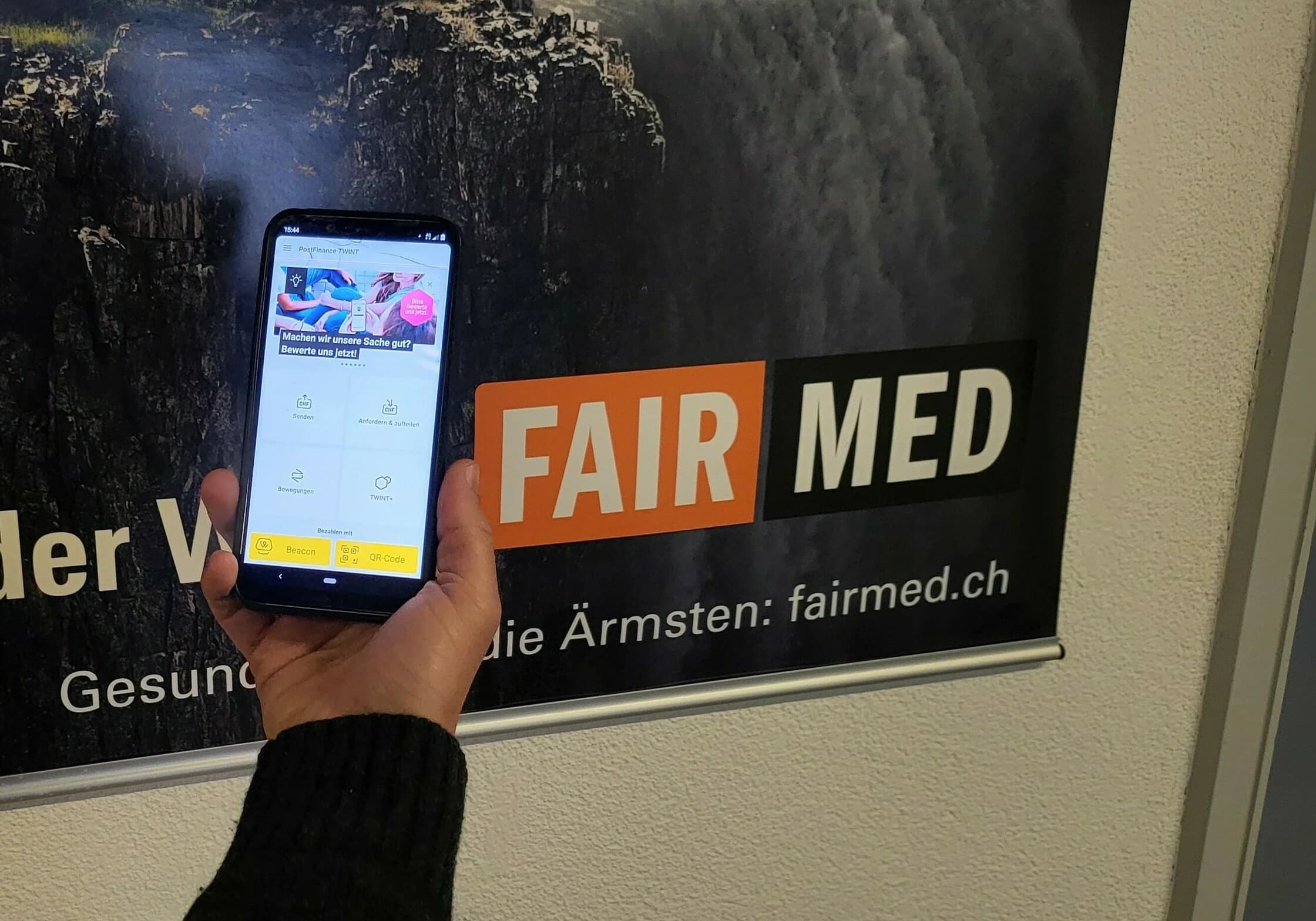 Auf dem Bild sieht man ein Handy, auf welchem die TWINT-App geöffnet ist. Im Hintergrund sieht man ein grosses FAIRMED-Plakat.