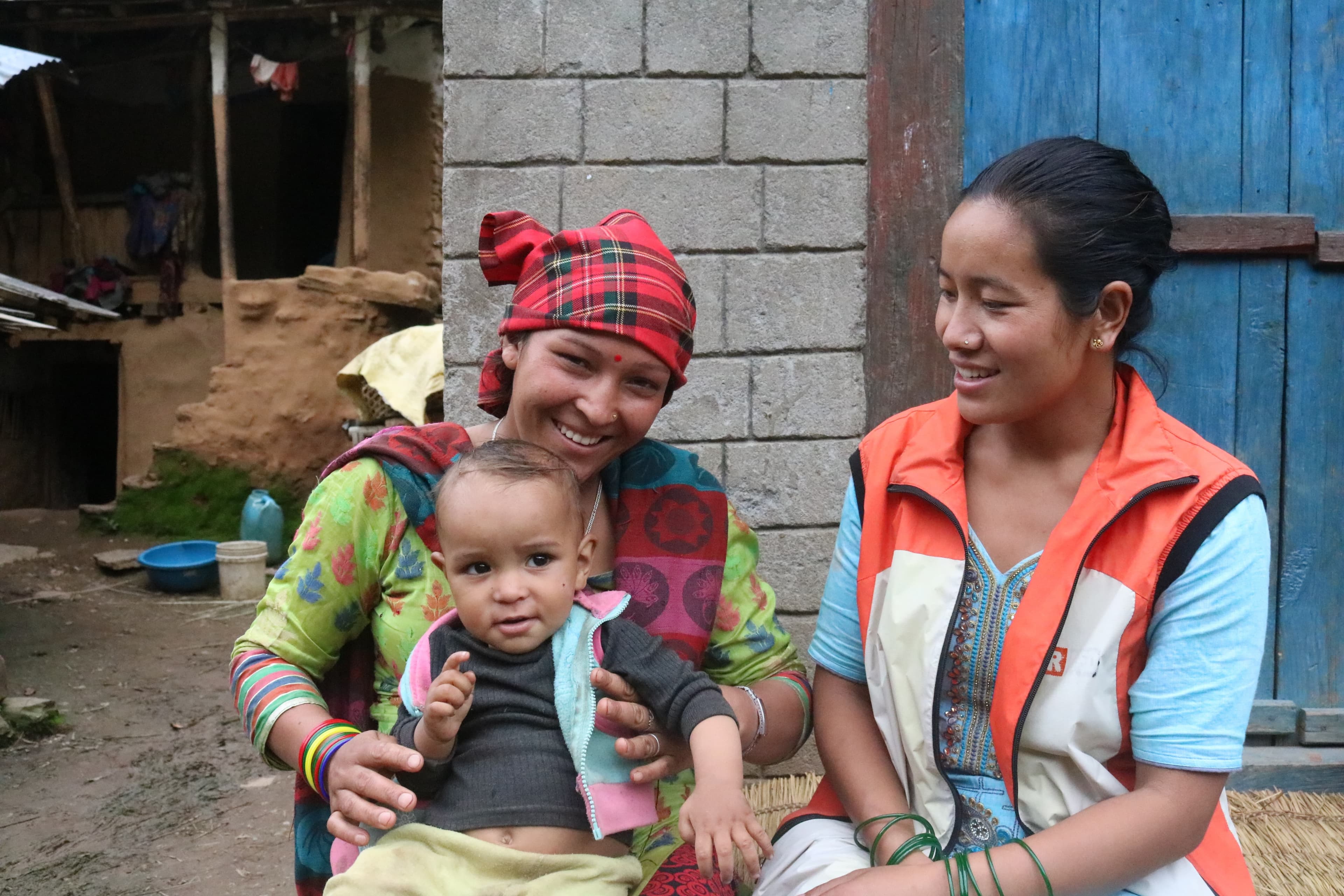 Auf dem Bild ist unsere Gesundheitshelferin Gyan zu sehen. Sie guckt ein Baby an, das auf dem Schoss seiner Mutter sitzt, und lächelt das Baby an. Alle drei sitzen im Freien vor einer Hütte und sind bunt gekleidet und lachen.