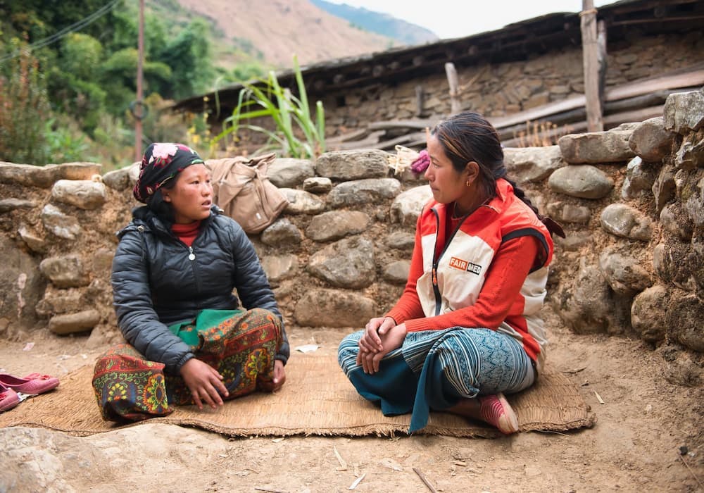 Gyan, a young FAIRMED employee, sits on the floor with a young mother and gives her advice about her health and the health of her child. They sit on a mat, with a stone wall and parts of a rudimentary hut in the background. The scene is set in a remote village in Baglung, Nepal