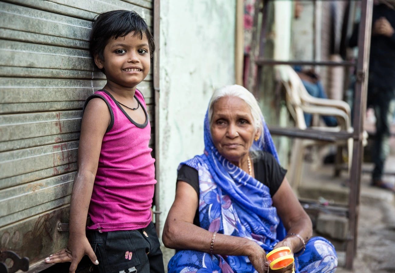 Ein kleines Mädchen steht gegen eine Wand gelehnt. Neben ihr sitz eine alte Frau in traditionellem indischen Gewand. Die Szenerie ist in einem Slum in Indien.