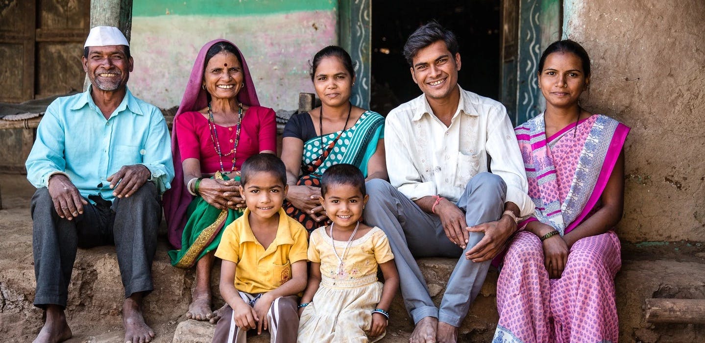 An Indian family sits outside in front of a house lined up for a photo. In the back are the grandparents, the parents, another person and in the foreground a girl and a boy.