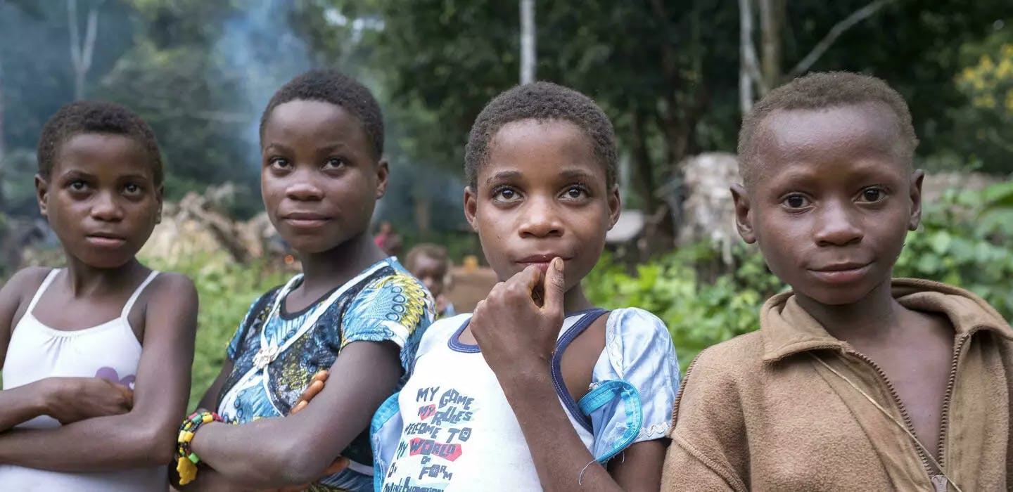 Vier Aka-Kinder aus unserem Projekt in der Zentralafrikanischen Republik stehe nebeneinander und lachen in die Kamera. Im Hintergrund ist der Dschungel zu erkennen, in dem ihr Dorf liegt.