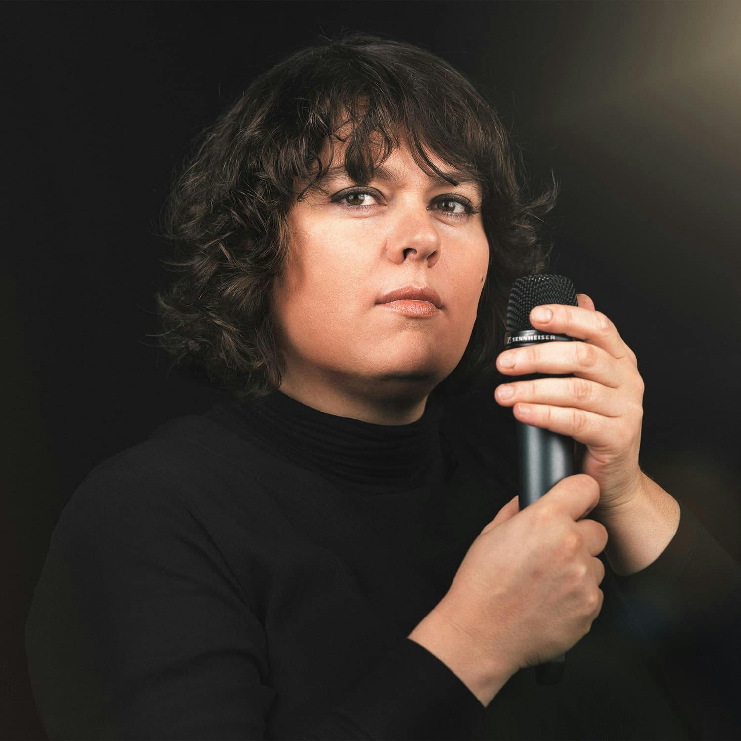 L'image montre Patti Basler devant un fond neutre, un micro à la main.
