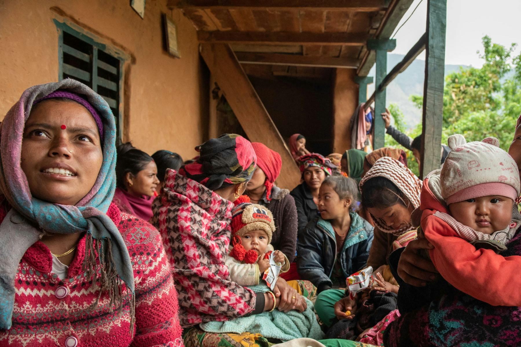The picture shows mothers and their children in Nepal. They are sitting close together under a roof. They are dressed in winter and colorful.