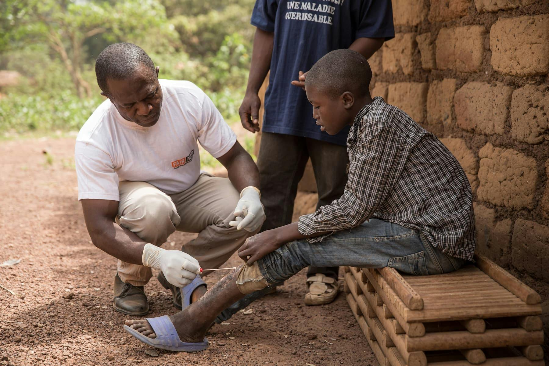 Un collaborateur de FAIRMED au Cameroun traite une blessure d'ulcère de Buruli à la jambe d'un adolescent. L'adolescent est assis devant une hutte dans un village reculé.