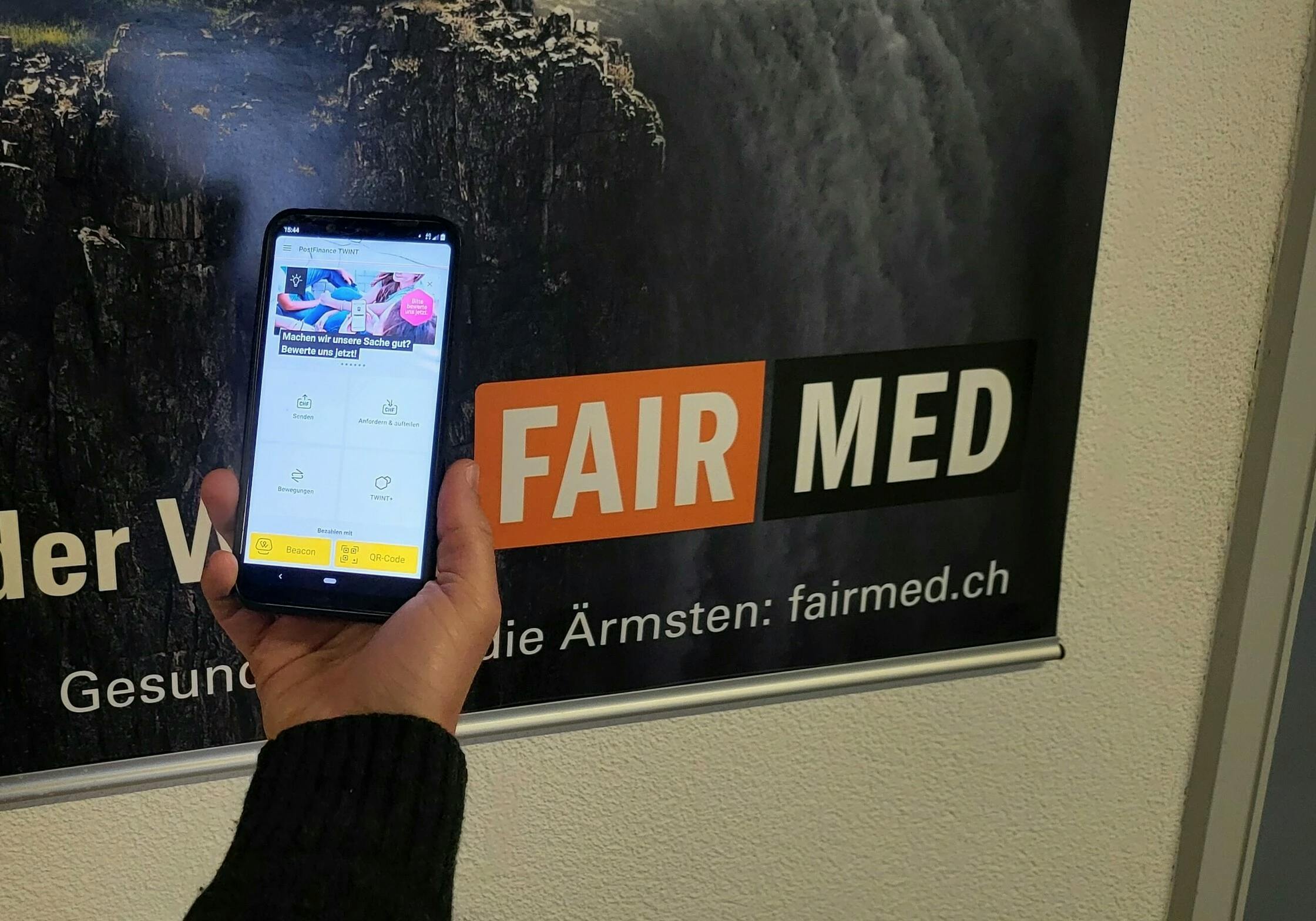 The picture shows a cell phone with the TWINT app open. In the background you can see a large FAIRMED poster.