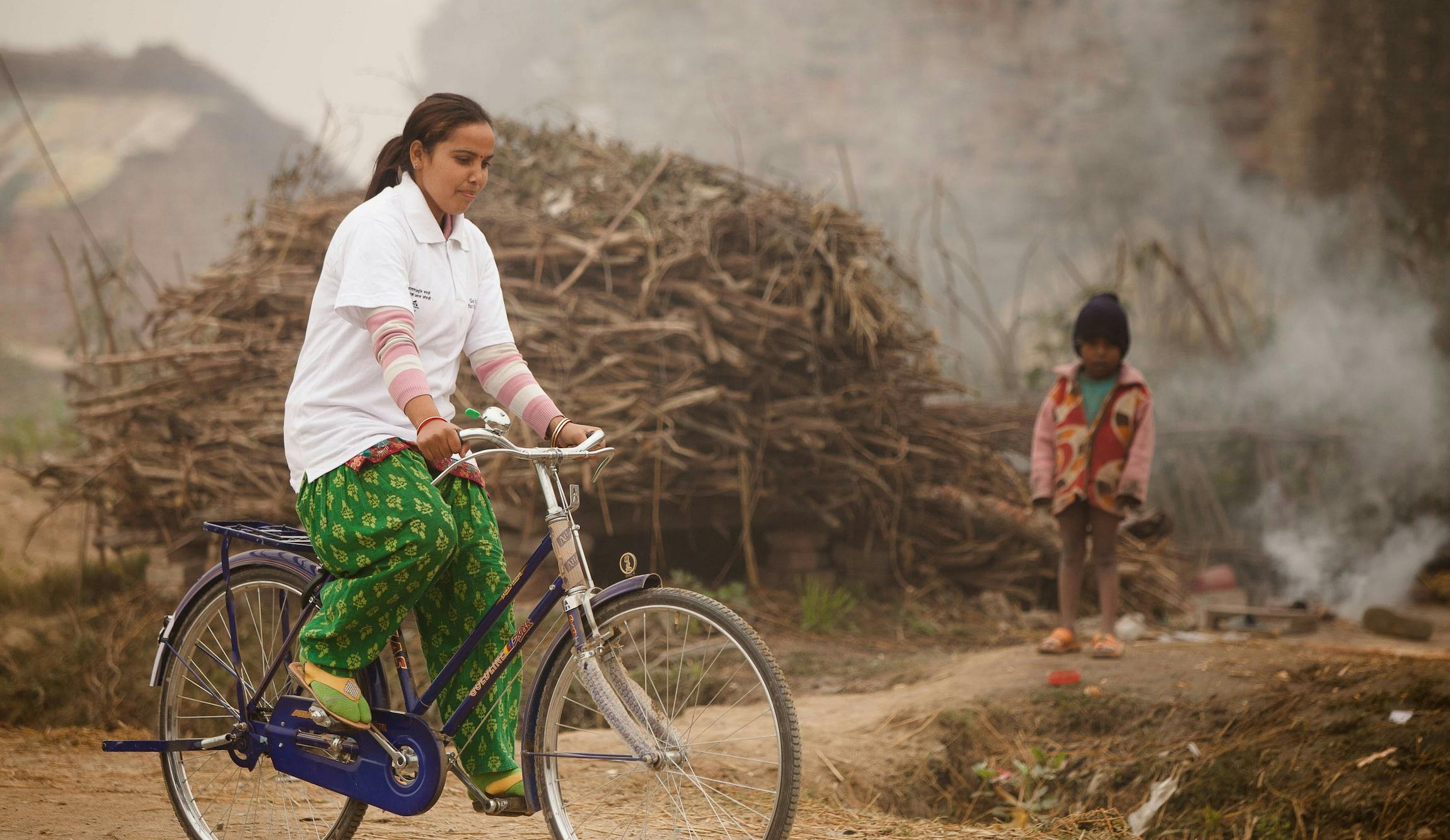 Eine FAIRMED-Mitarbeiterin, gekleidet in grünen Hosen und einem weissen Polo-Shirt mit FAIRMED-Logo fährt mit einem Velo. Im Hintergrund ist eine karge Landschaft in Nepal zu sehen. Ausserdem ist im Hintergrund ein Mädchen zusehen, das eher winterlich gekleidet ist.