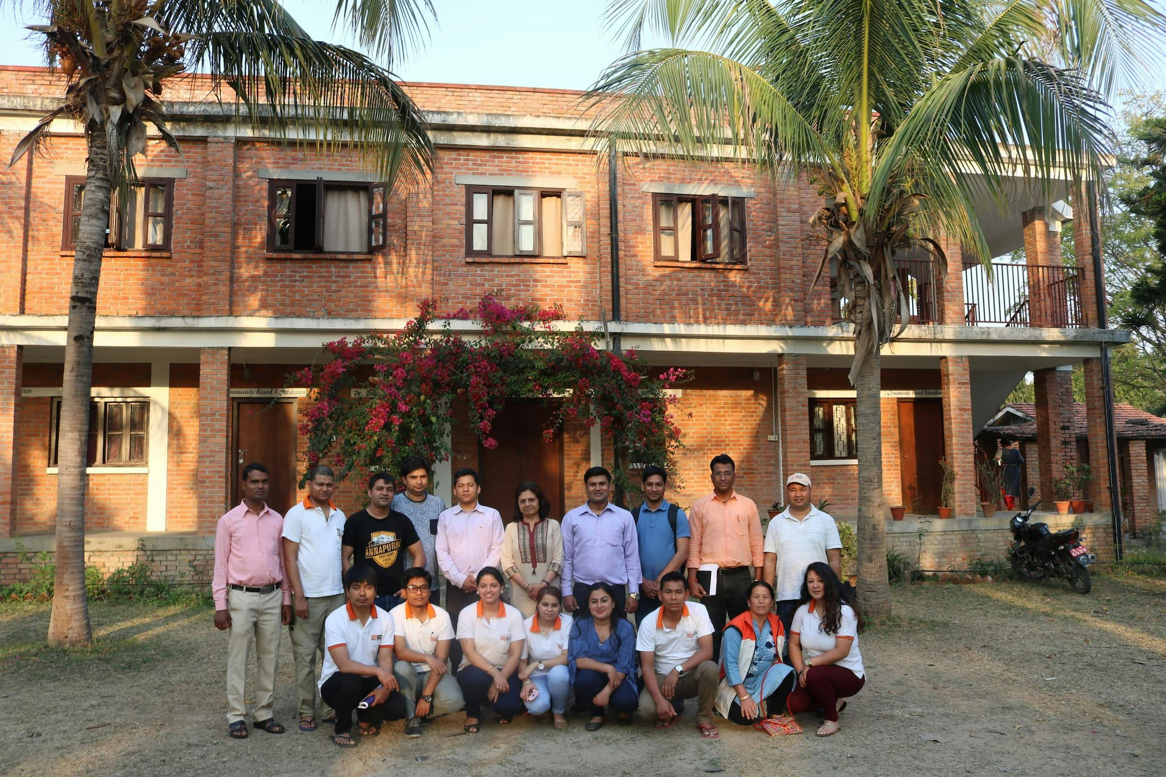 A group photo of the Nepal team. Some of them are wearing a white polo shirt with orange collar and FAIRMED logo. The group is lined up outside in front of the FAIRMED office in Nepal. It is warm and palm trees can be seen.
