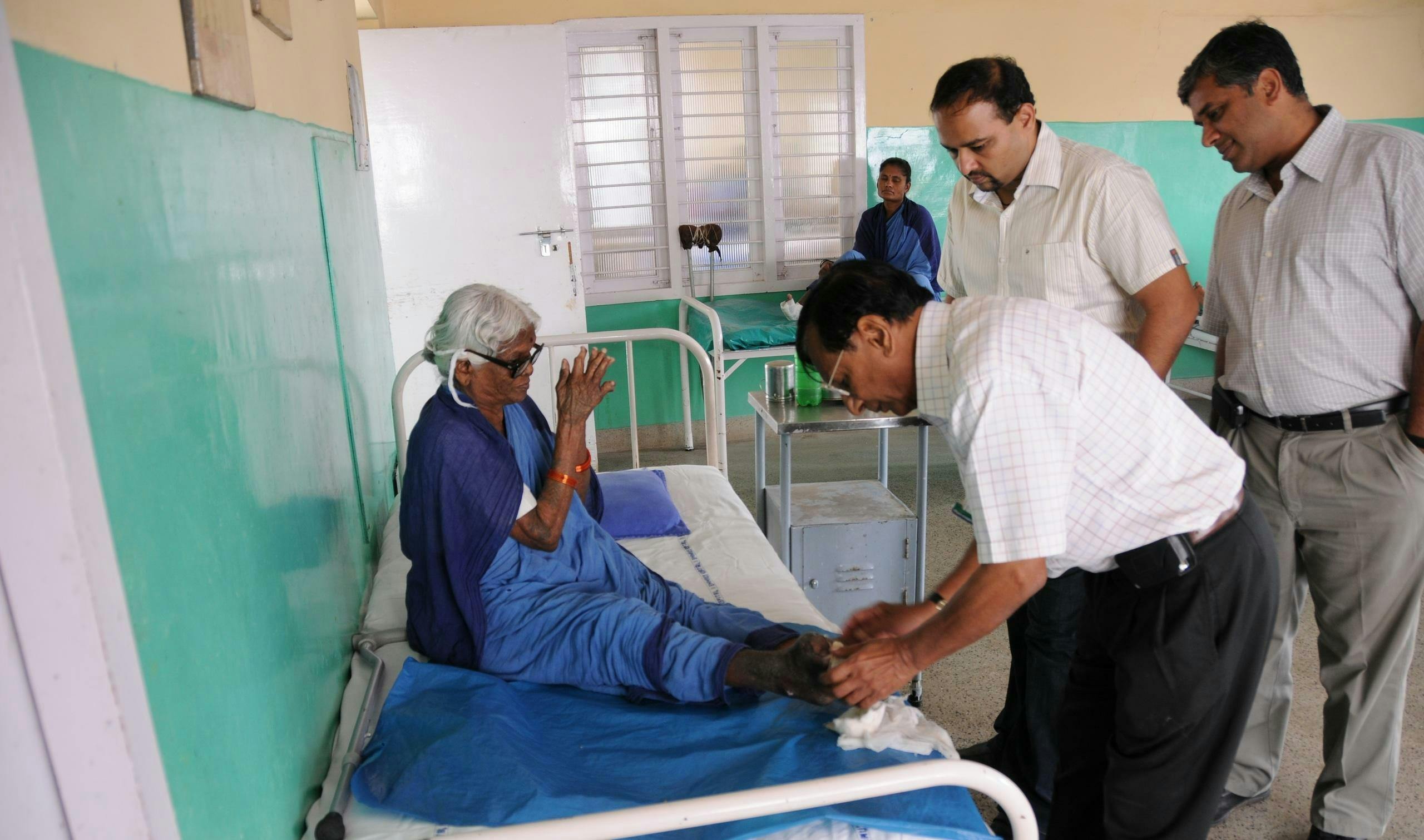 Une femme âgée est assise sur un lit d'hôpital dans un hôpital indien et joint ses mains. Un médecin examine son pied à la recherche de signes de lèpre. Deux hommes se tiennent derrière et observent.