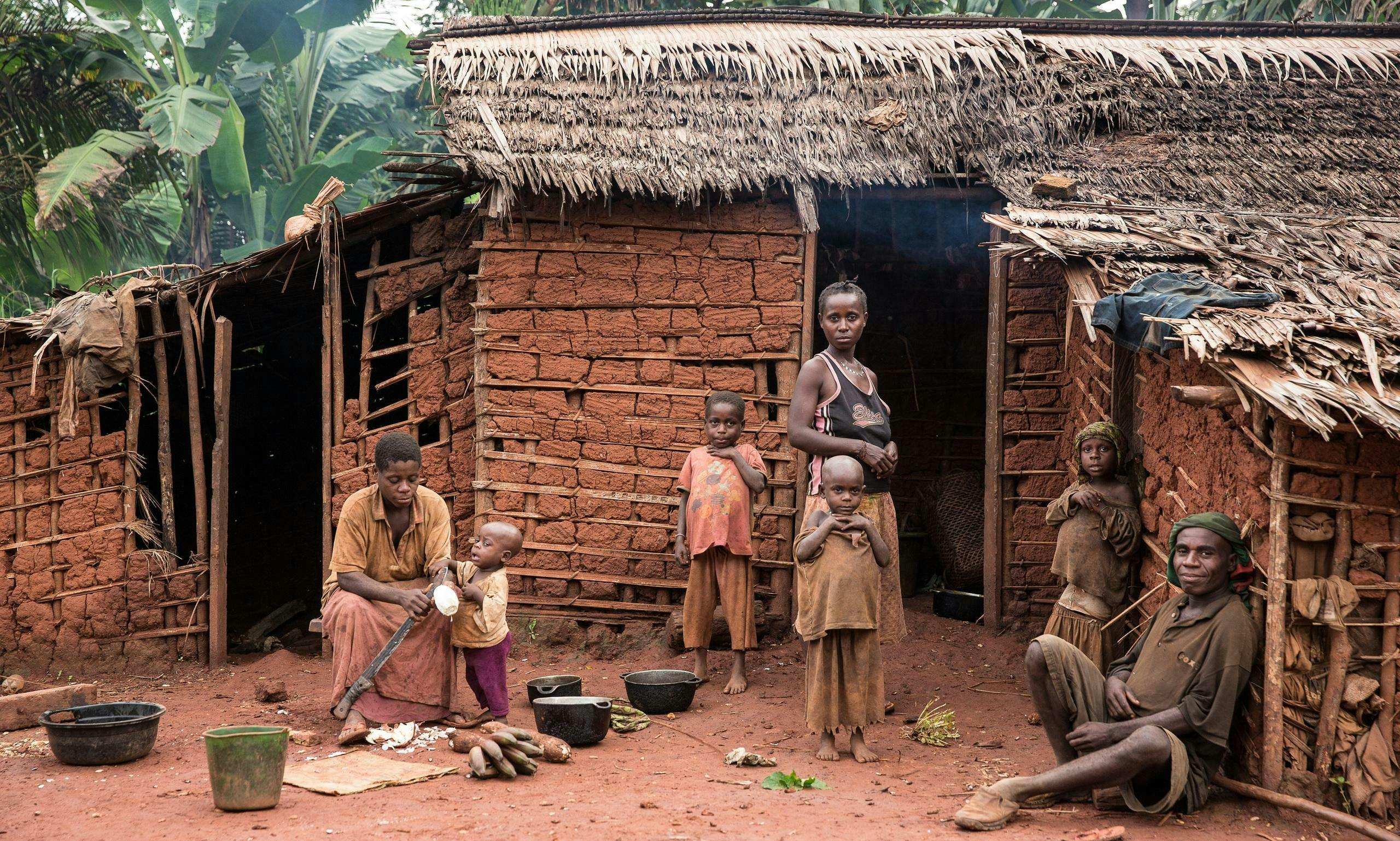 A Baka family in front of a mud hut in their village. A mother and her child are sitting on the ground preparing food. Three other children and a young woman stand in front of the entrance. An elderly man sits on the ground and leans against a wall.