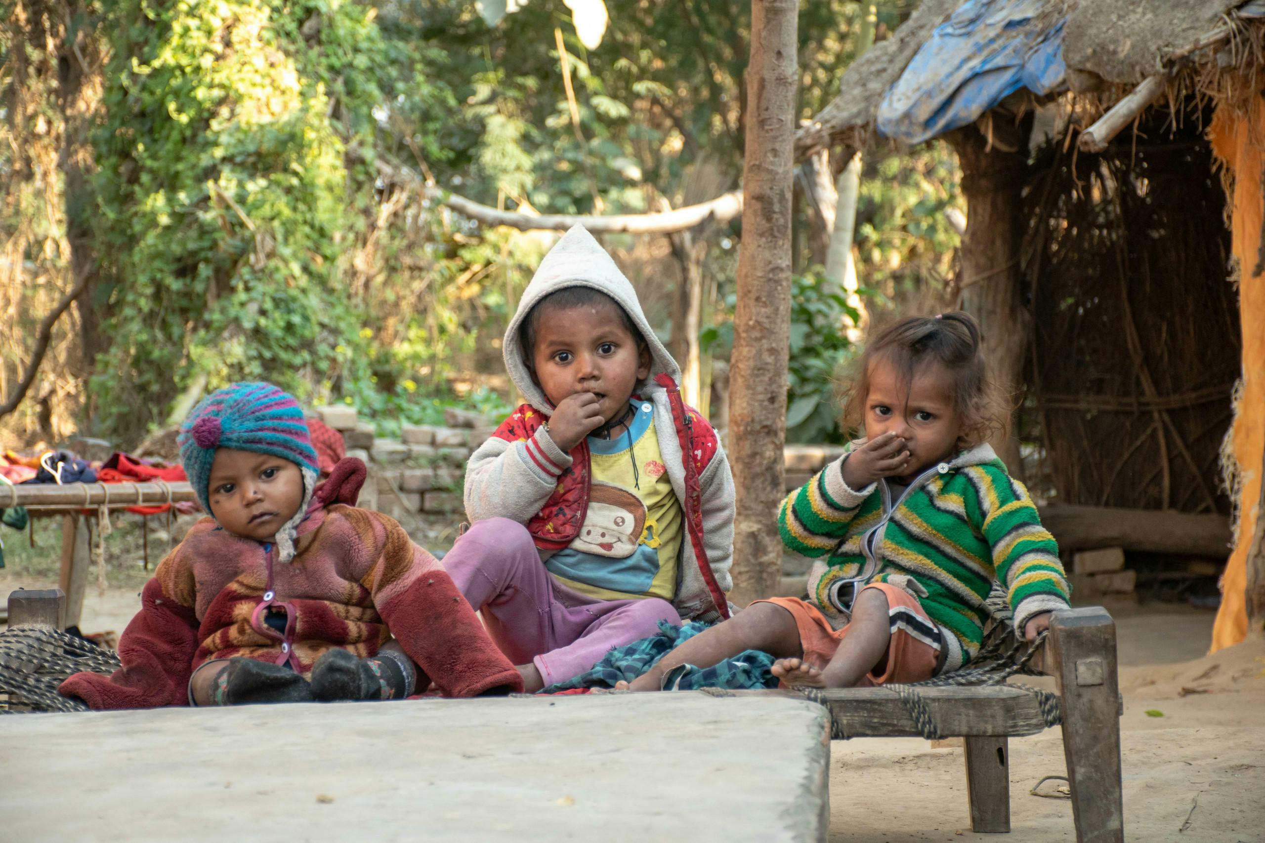 Three small children from Nepal look into the camera. They are dressed in winter clothes. In the background you can see part of a rudimentary hut and the forest of the project area, which is far away.