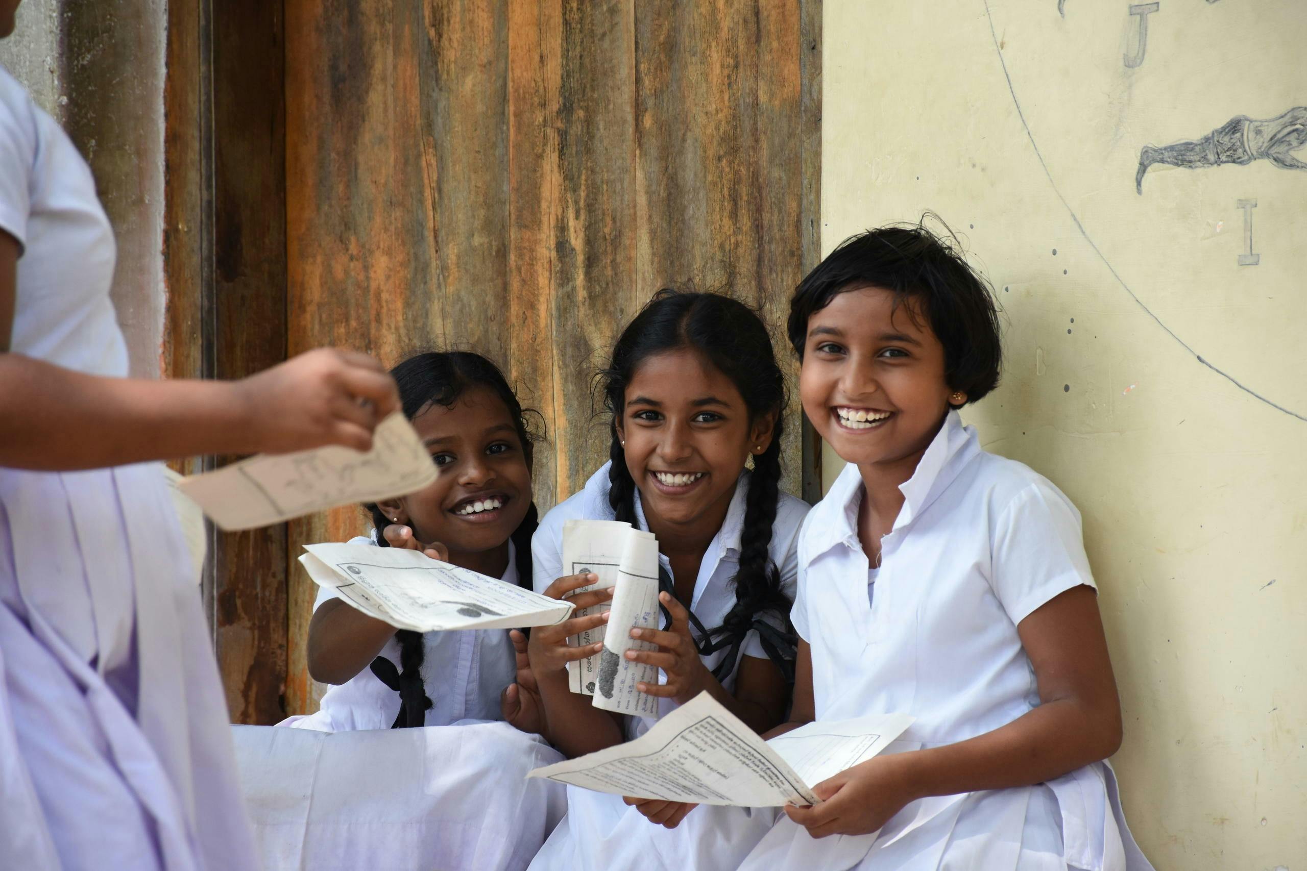 Drei lachende Mädchen in ihrer typischen weissen Schuluniform lachen in die Kamera. Sie halten Papier in den Händen und sitzen im Freien an einer Wand.