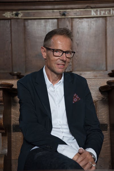 Bänz Friedli se tient devant un fond neutre. Il porte une chemise blanche, un blazer noir, une pochette colorée et des lunettes noires.
