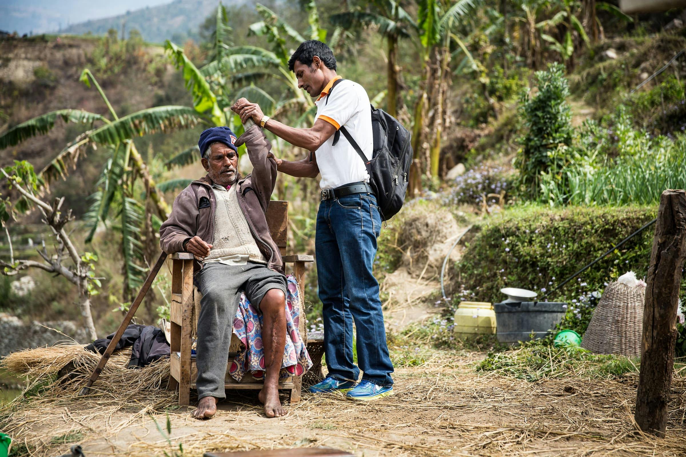 A young FAIRMED worker stretches the arm of an old man to see if his leprosy disease affects his mobility. The old man is sitting on a chair outside. Palm trees and trees can be seen in the background. The scenery is set in rural India.