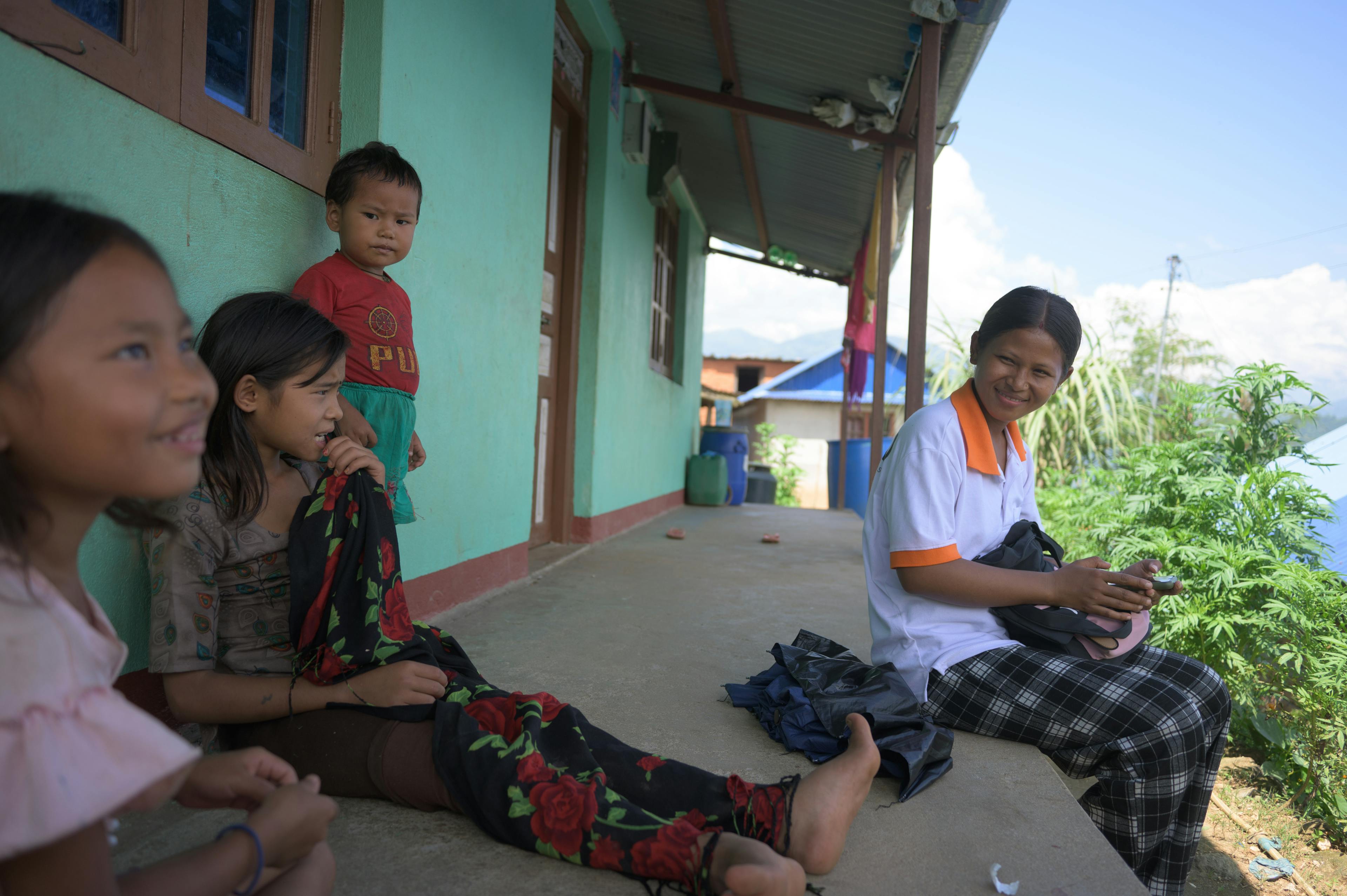 Sujita sitzt mit drei kleinen Kindern auf der Veranda vor einem Haus. Sie trägt ein FAIRMED-Shirt und karierte Hosen.