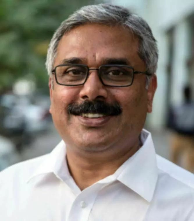 Our country representative John Kurian George. He wears a white shirt, a black moustache and black glasses. His hair is gray.