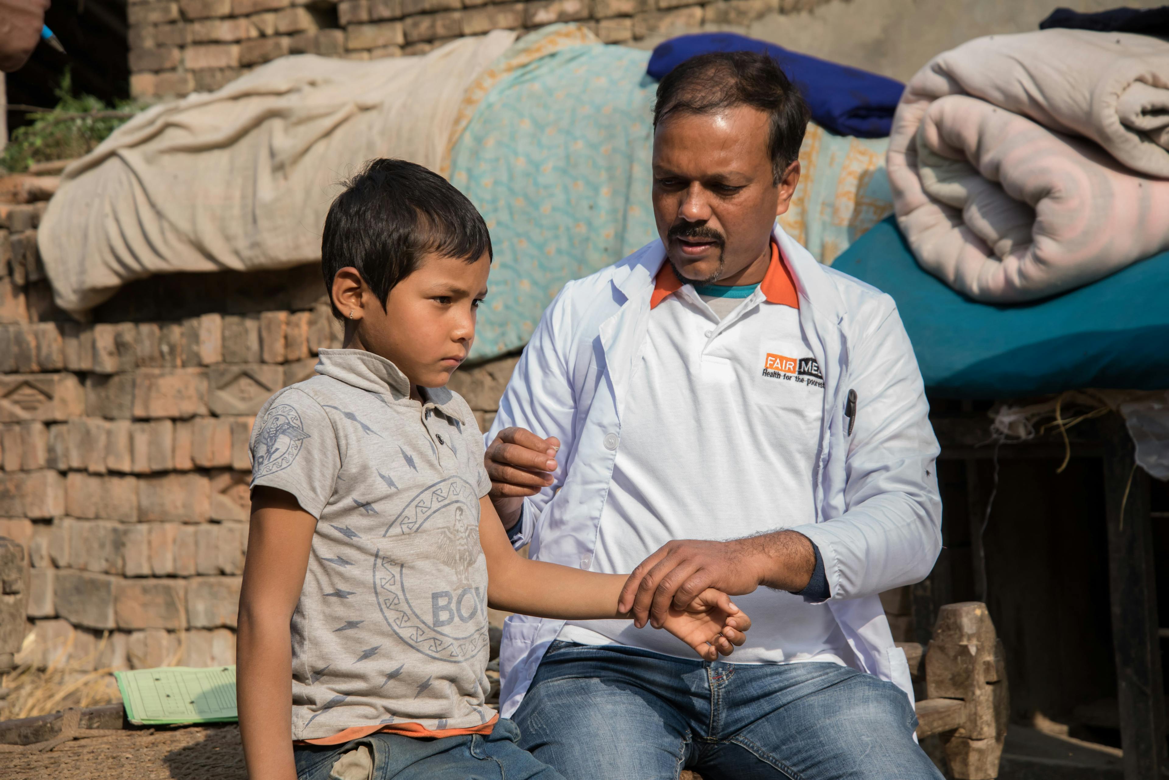 FAIRMED Mitarbeiter untersucht Kind in Indien. Sie sitzen gemeinsam auf einer Mauer und der FAIRMED-Mitarbeitende untersucht den Arm des Knaben.