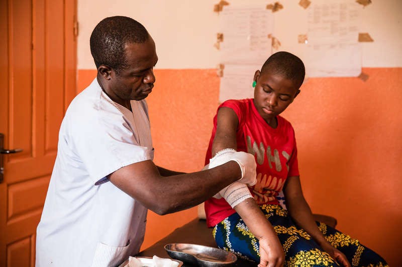 Une fillette avec une blessure d'ulcère de Buruli au bras est soignée par un collaborateur de FAIRMED. La jeune fille est assise sur une table d'examen dans un hôpital de Bankim, au Cameroun. La salle de soins est très rudimentaire.