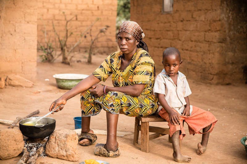A mother without toes sits on a stool and cooks something over a fireplace. She is colorfully dressed, next to her sits her son, who also has feet mutilated by leprosy. In the background the walls of mud huts can be seen.
