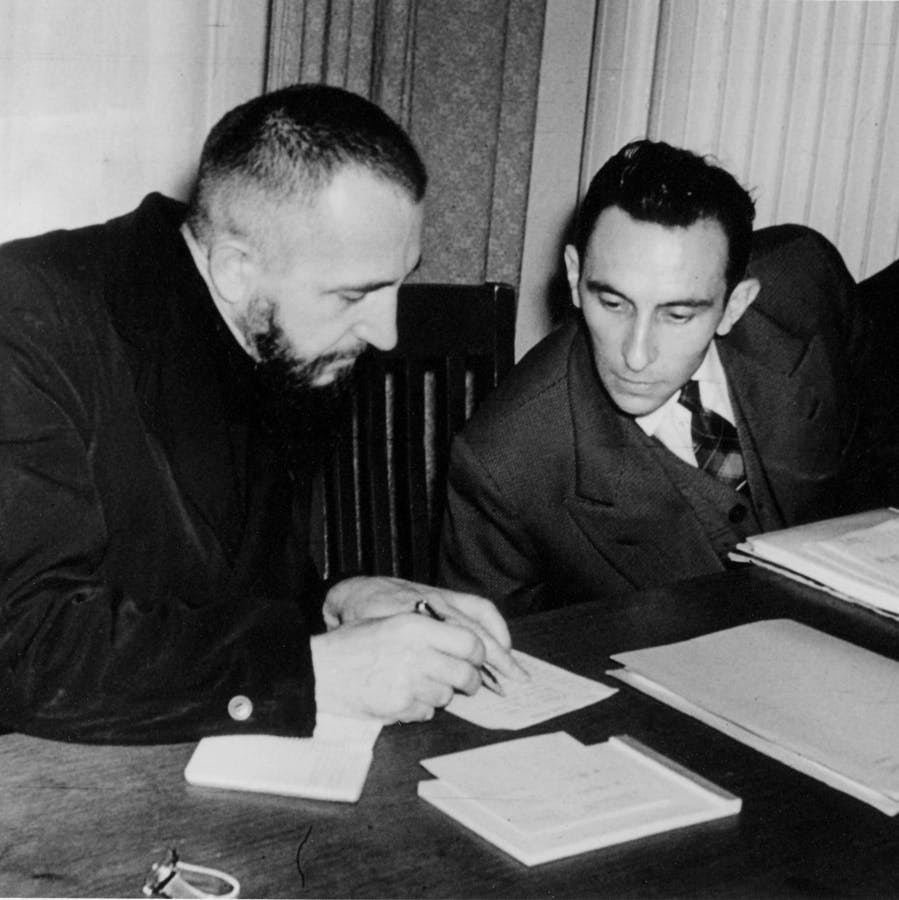 A historical black and white image showing Abbé Pierre and Marcel Farine sitting at a desk discussing documents.