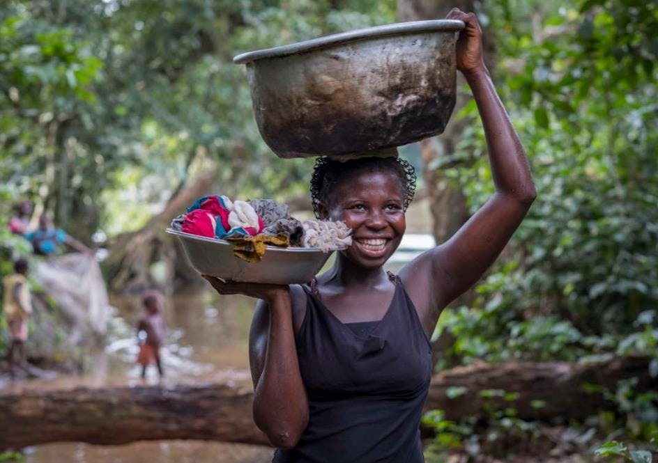 The picture shows a young woman carrying on her head a trough with water in her left hand and a container with laundry in her right hand. In the background there is a body of water in the middle of the jungle and some trees and a bathing child.