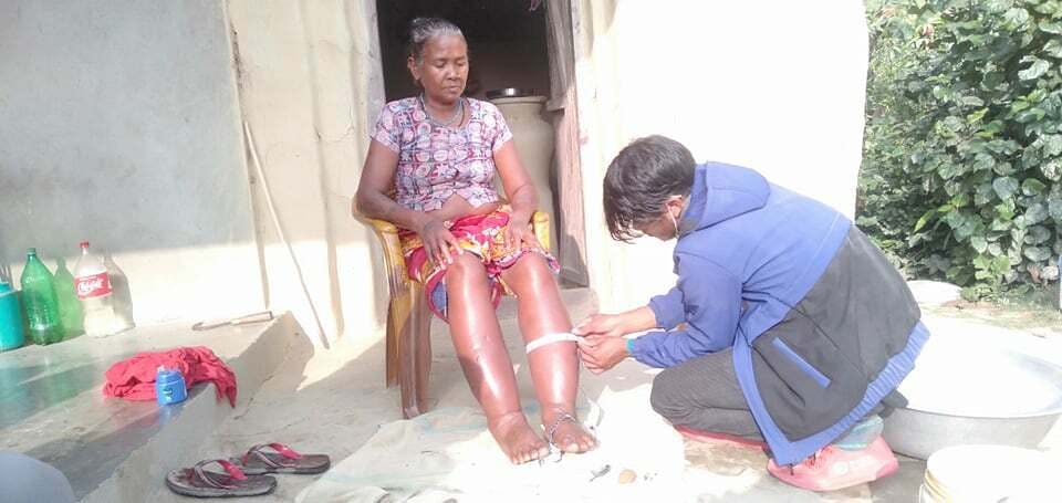 La photo montre Soharatiya Tharu assise sur une chaise devant sa maison. Un agent de santé mesure la circonférence de ses jambes.